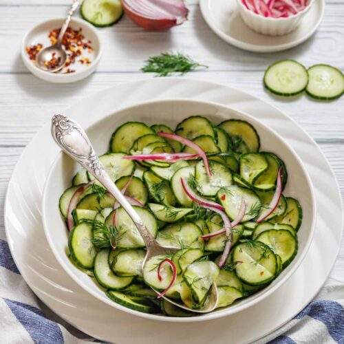A bowl of Quick Marinated Cucumbers with red onion and dill, served with a spoon. Sliced cucumbers, onions, and seasoning are visible on the table nearby.