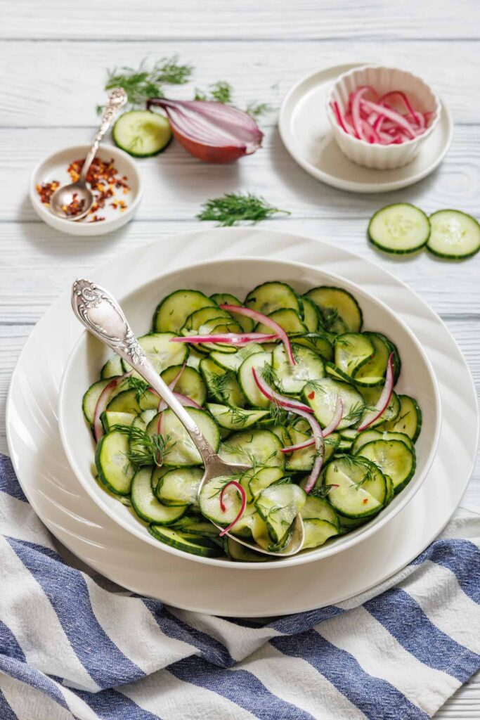 A bowl of Quick Marinated Cucumbers with red onion and dill, served with a spoon. Sliced cucumbers, onions, and seasoning are visible on the table nearby.