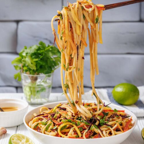 A bowl of vegetable noodle stir-fry with chopsticks lifting noodles, garnished with herbs; lime, cilantro, and dipping sauce are in the background.