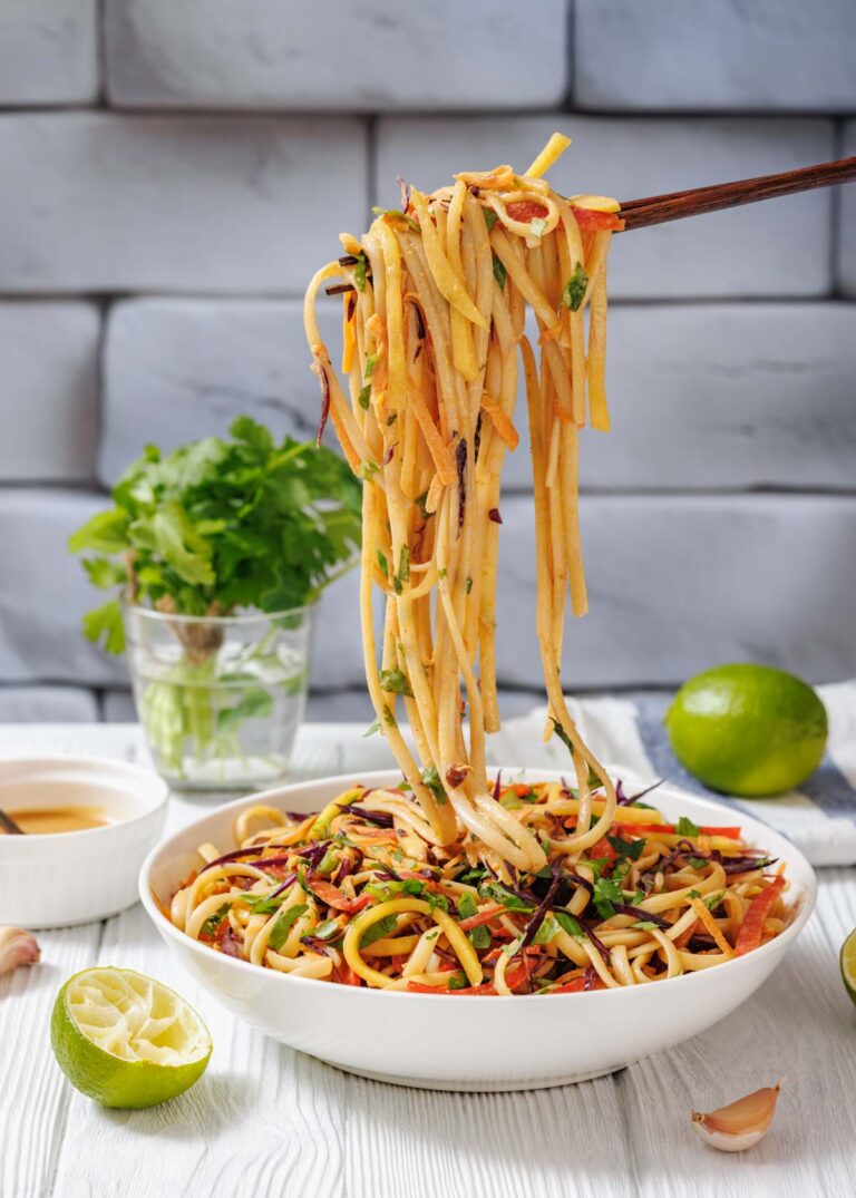 A bowl of vegetable noodle stir-fry with chopsticks lifting noodles, garnished with herbs; lime, cilantro, and dipping sauce are in the background.