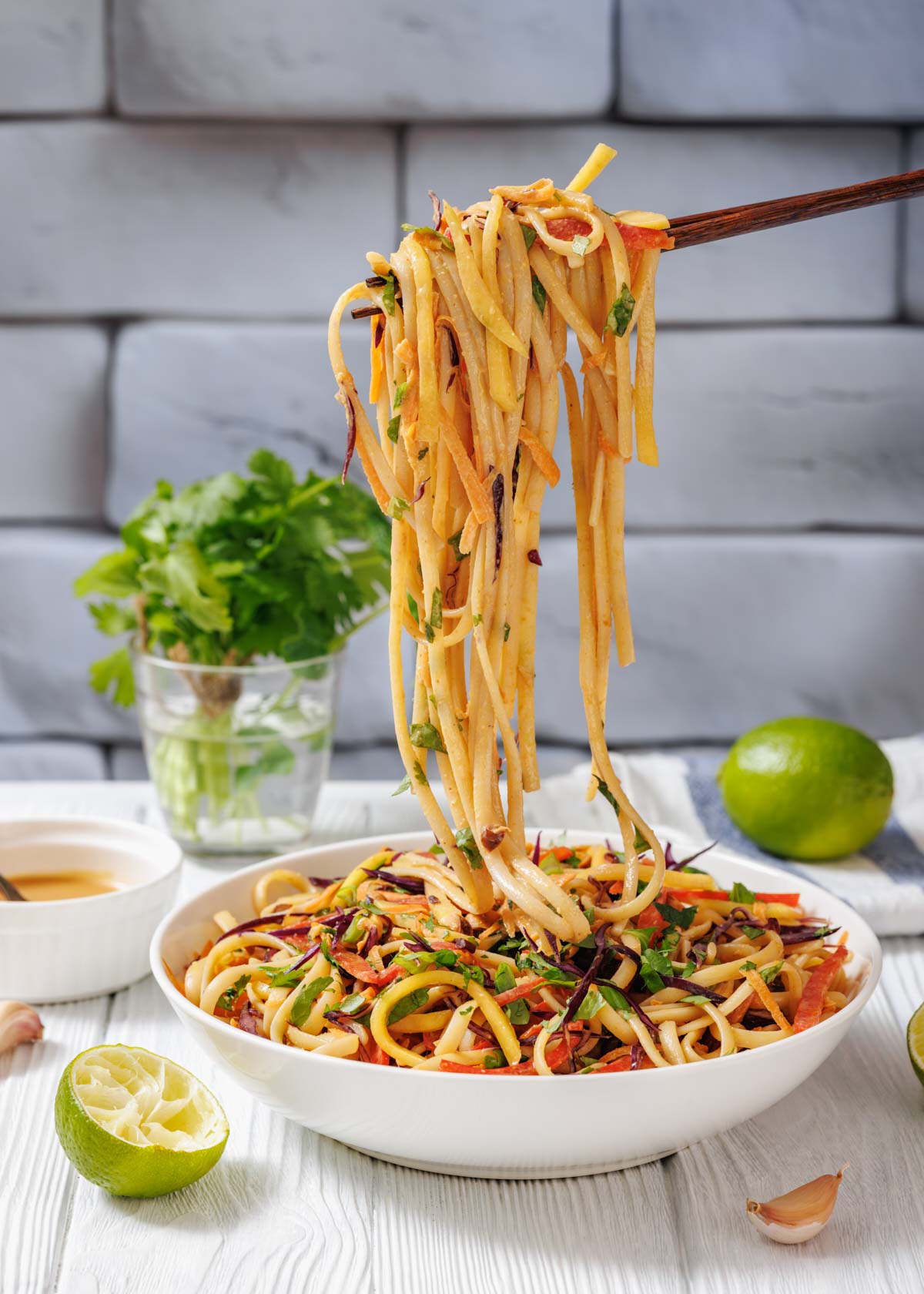 A bowl of vegetable noodle stir-fry with chopsticks lifting noodles, garnished with herbs; lime, cilantro, and dipping sauce are in the background.