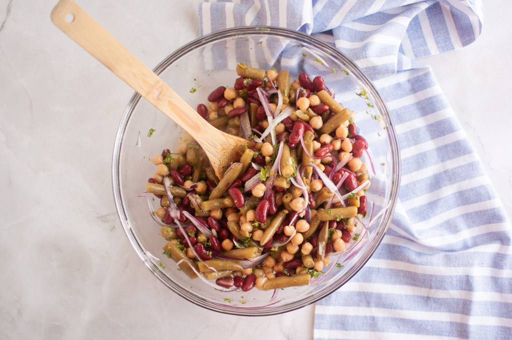 A glass bowl filled with a Three Bean Salad including kidney beans, chickpeas, green beans, and sliced red onion, with a wooden spoon and a striped cloth nearby.