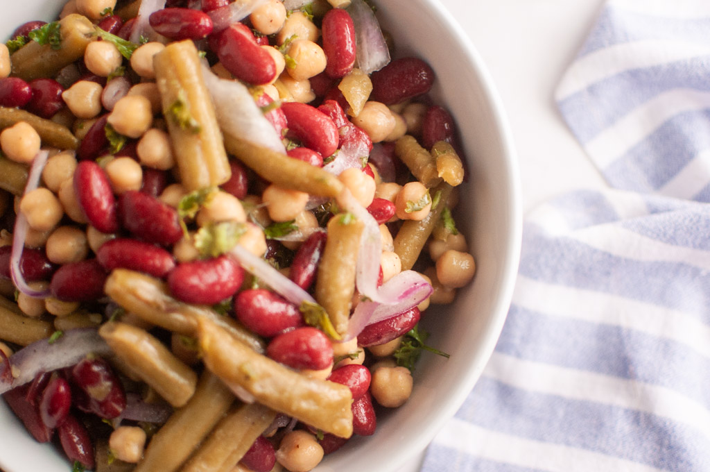 A bowl of mixed bean salad with kidney beans, chickpeas, green beans, and sliced red onions, next to a blue and white striped cloth.