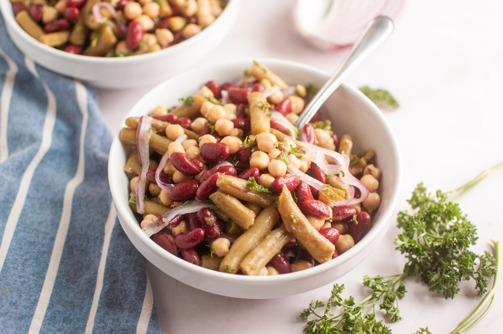 Bowl of ClassicThree Bean Salad featuring kidney beans, chickpeas, and green beans, garnished with thinly sliced red onions and fresh parsley.