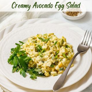 A white plate with avocado egg salad, garnished with herbs, is shown with a fork, cloth, and a small bowl of spices.