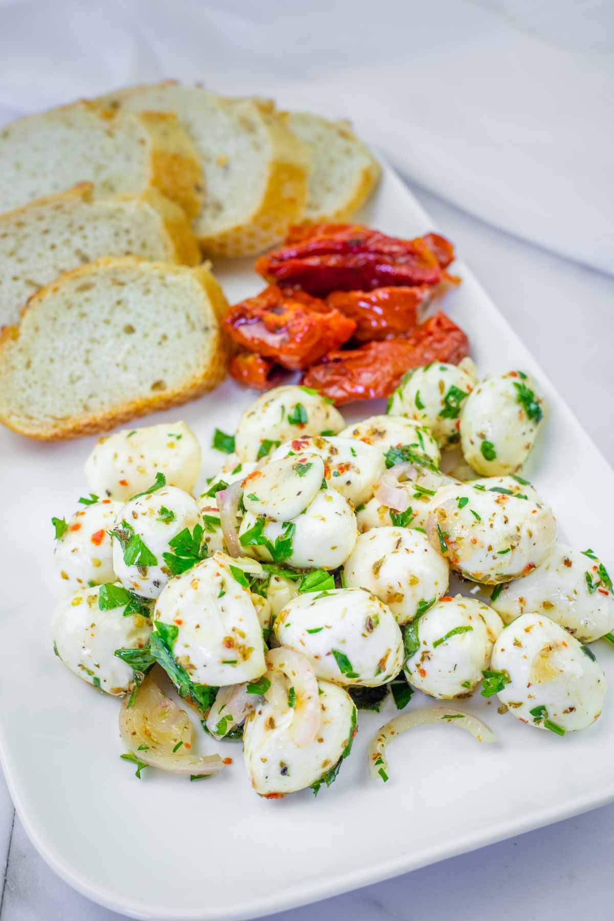 A rectangular plate with sliced bread, sun-dried tomatoes, and marinated mozzarella balls garnished with herbs and onions.