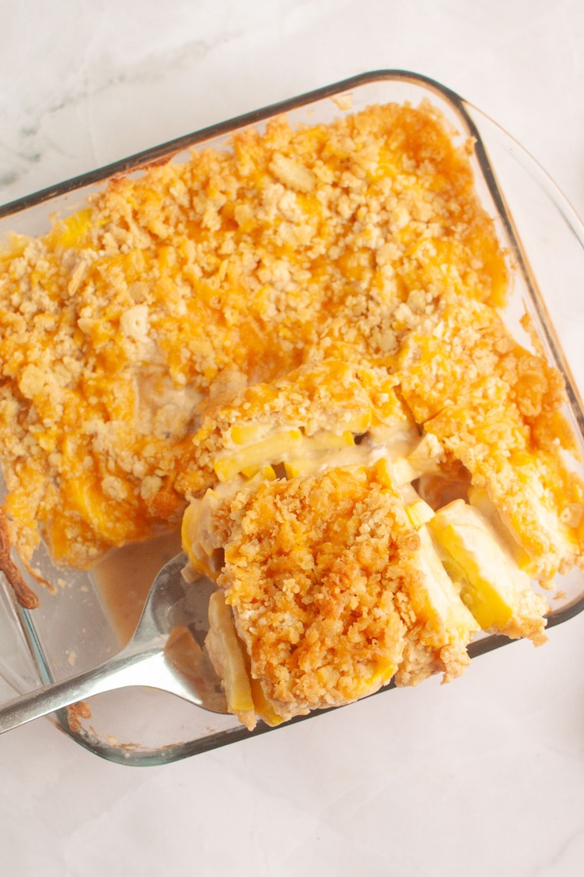 A glass baking dish with a serving of cheesy squash casserole topped with a golden crumb crust, partially scooped out with a metal spatula.