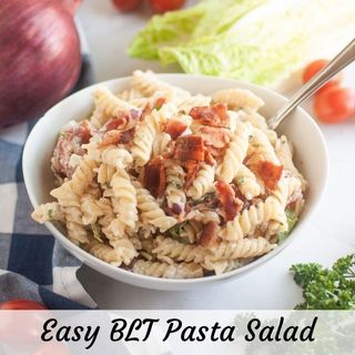 A white bowl filled with BLT pasta salad, featuring spiral pasta, creamy dressing, pieces of bacon, and other visible ingredients, is shown with a spoon. Red onions, lettuce, cherry tomatoes, and parsley are in the background.