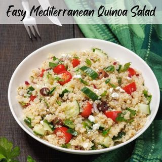 A bowl of quinoa salad with tomatoes, cucumbers, olives, and feta cheese. A fork and knife are beside the bowl on a wooden table with a green plaid cloth.
