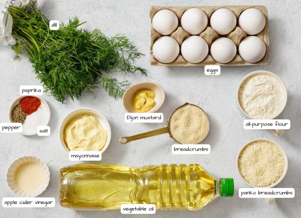 Overhead shot of deviled egg ingredients, including eggs, dill, spices, mayonnaise, mustard, breadcrumbs, flour, and oil, arranged on a light surface with labels for each ingredient.