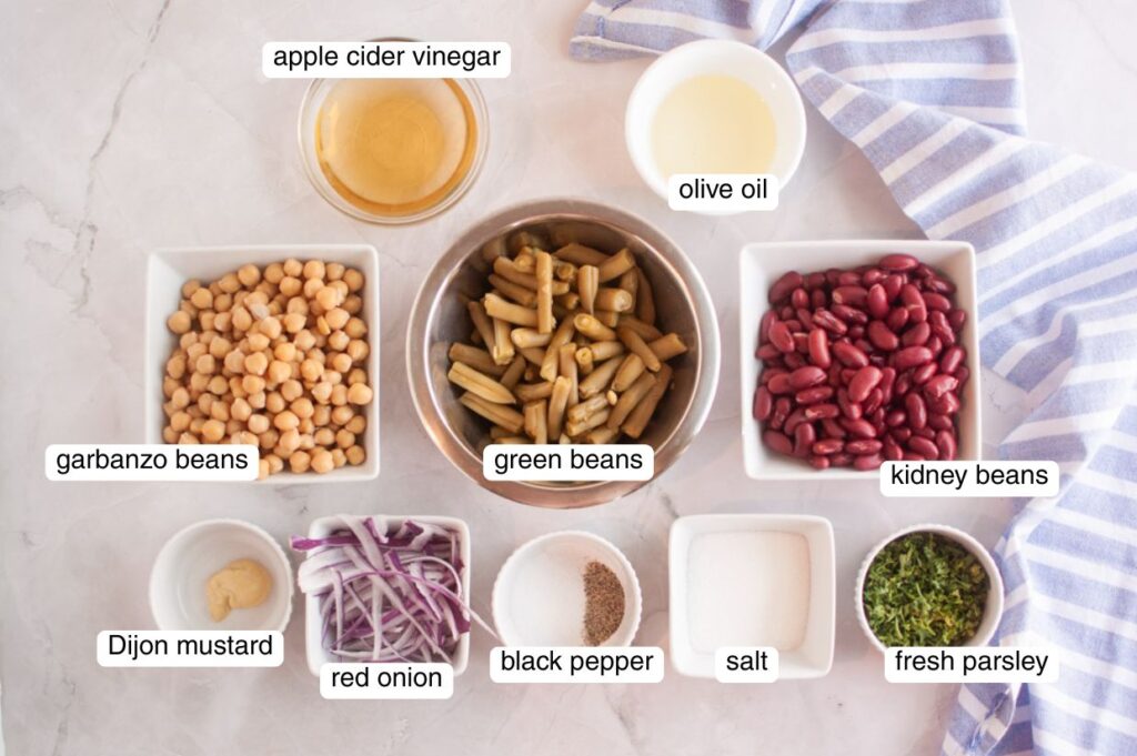 Overhead view of labeled ingredients for Grandma&rsquo;s Three Bean Salad, including garbanzo beans, green beans, kidney beans, red onion, Dijon mustard, black pepper, salt, fresh parsley, apple cider vinegar, and olive oil arranged on a light surface with a striped cloth.