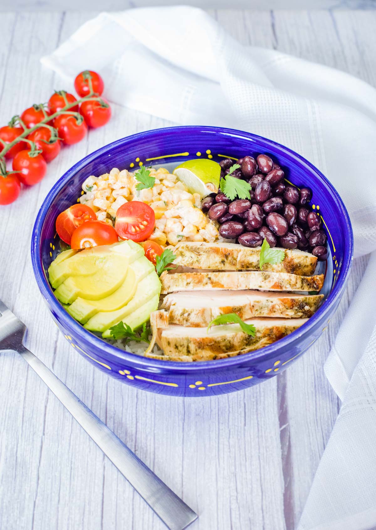 A blue bowl filled with a street corn chicken rice bowl, featuring grilled chicken, corn, tomatoes, avocado, and black beans, is shown with a fork, cloth, and tomatoes.