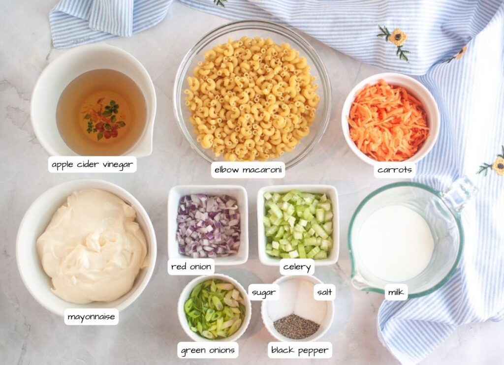 Overhead shot of ingredients for Hawaiian macaroni salad, including macaroni, carrots, mayonnaise, onions, celery, milk, seasonings, and vinegar, arranged on a light surface with a striped cloth and labels for each ingredient.