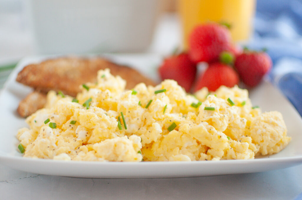 A plate of scrambled eggs garnished with chives, served with toast and fresh strawberries, with a glass of orange juice in the background.