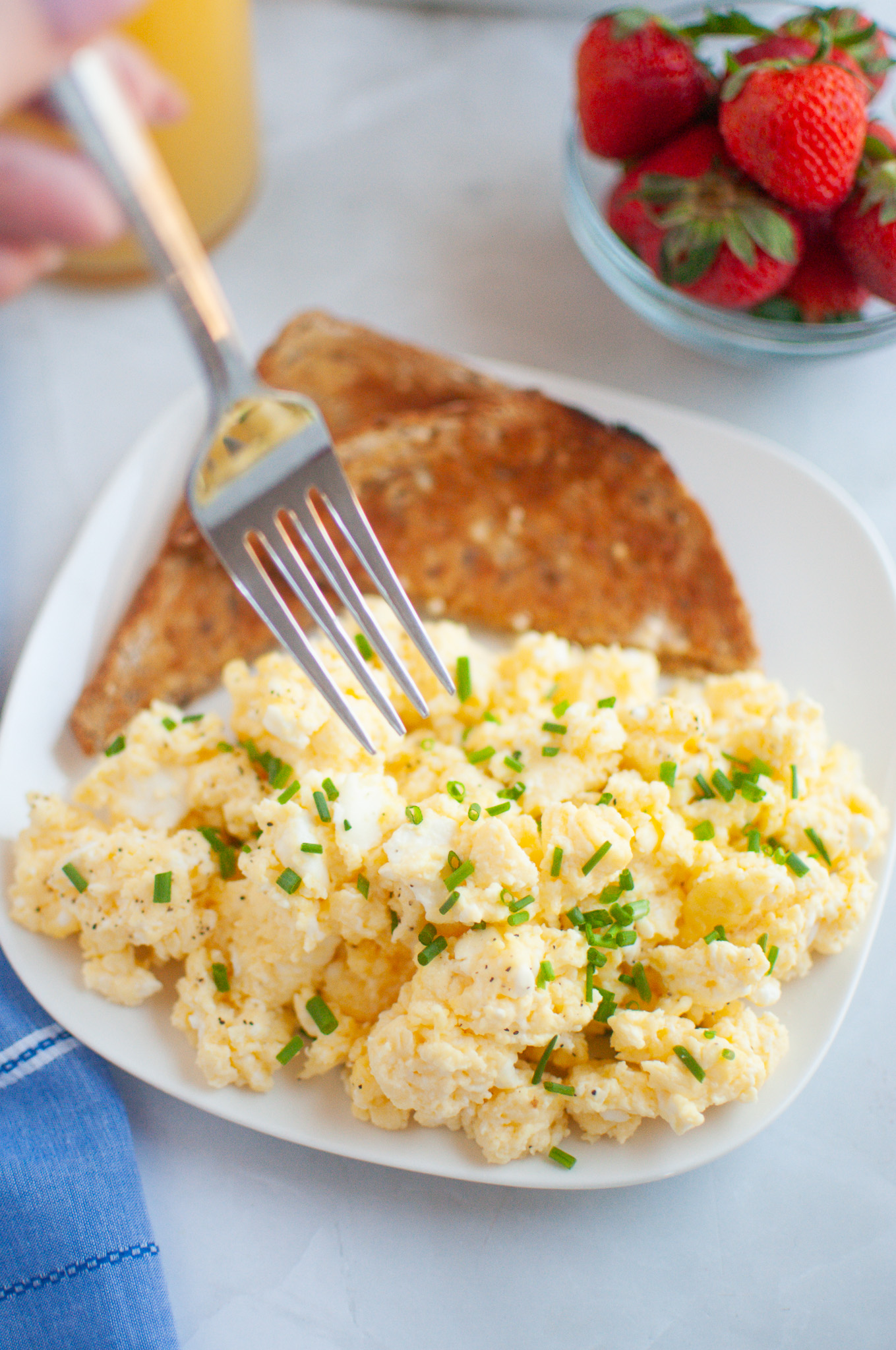 A plate of scrambled eggs with chives and toast, with a fork taking a bite, alongside a glass of orange juice and a bowl of strawberries.