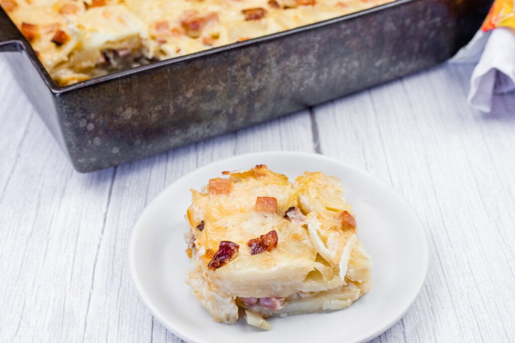 A slice of Scalloped Potatoes with Ham and Cheese is served on a white plate, with the rest of the dish visible in a metal baking pan on a light wooden surface.