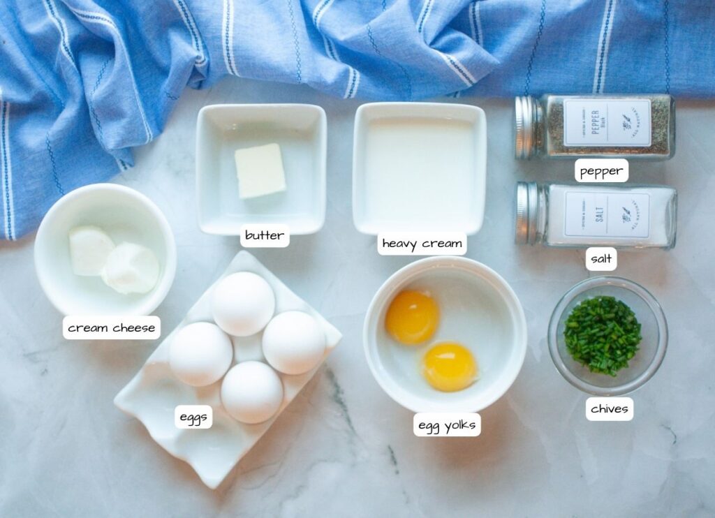 Top-down view of ingredients on a countertop: cream cheese, butter, heavy cream, four eggs, two egg yolks, chopped chives, salt, and pepper, with a blue cloth in the background.