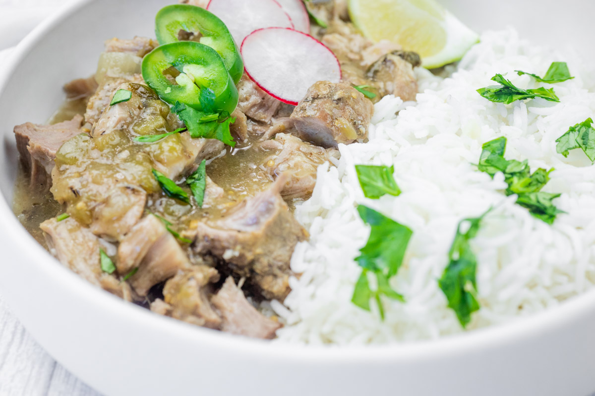 Close-up of a bowl of slow cooker pork chile verde served with white rice, garnished with fresh cilantro, sliced jalape&ntilde;os, radish slices, and a wedge of lime.