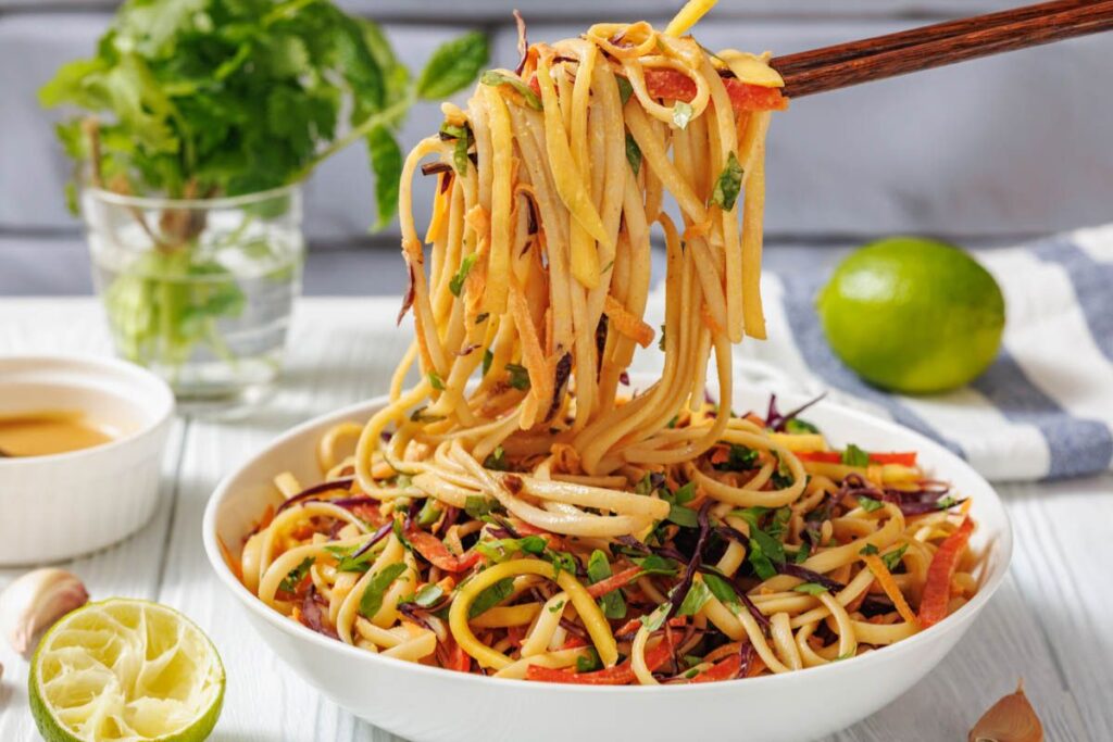 A bowl of vegetable noodle salad with chopsticks lifting a portion; fresh herbs, lime, and sauce are in the background.