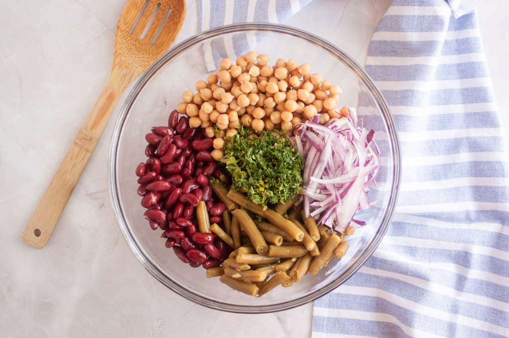 Overhead view of ingredients for Three Bean Salad, including garbanzo beans, green beans, kidney beans, red onion, Dijon mustard, black pepper, salt, fresh parsley, apple cider vinegar, and olive oil arranged on one bowl with a striped cloth.