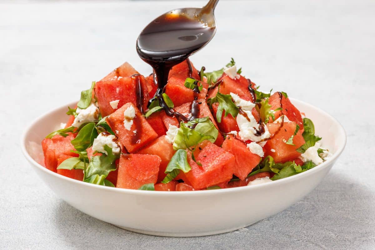 A spoon drizzles balsamic glaze over a salad with watermelon cubes, crumbled feta, and arugula in a white bowl on a light surface.