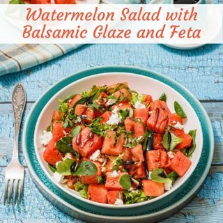 A bowl of watermelon salad topped with feta, fresh herbs, and balsamic glaze sits on a blue wooden table with a fork and napkin beside it.
