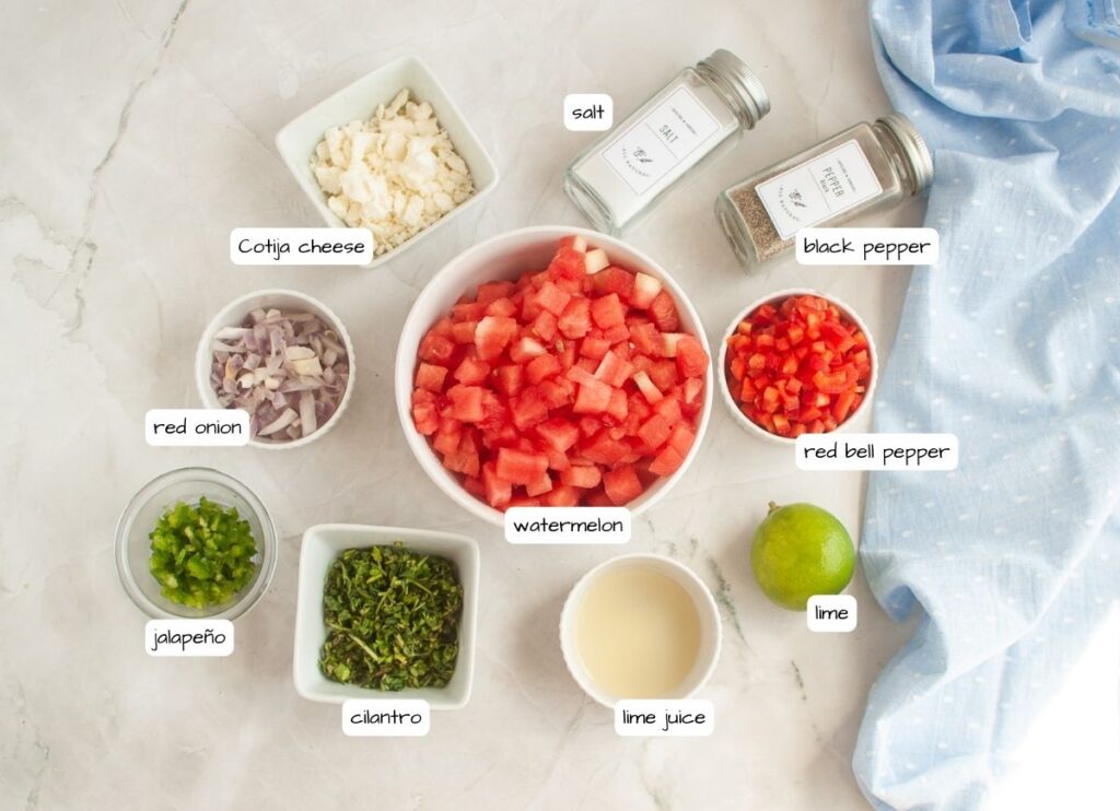 Overhead shot of ingredients for watermelon salad laid out in small bowls on a white marble surface, including diced watermelon, red bell pepper, shallots, feta cheese, jalapeno, cilantro, lime juice, lime, salt, and pepper. A blue cloth is draped on the side.