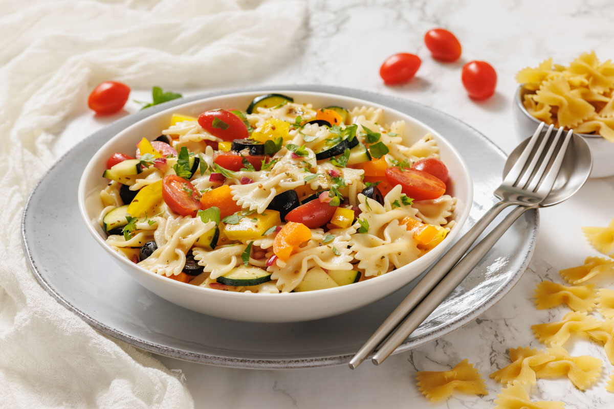 Bowl of bowtie pasta salad with cherry tomatoes, zucchini, bell peppers, and herbs, served on a plate with a fork and spoon.