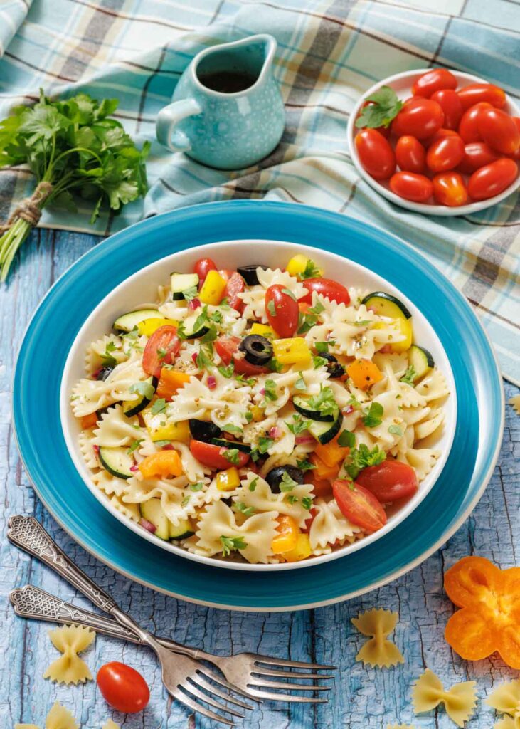 A bowl of farfalle pasta salad with cherry tomatoes, bell peppers, olives, zucchini, red onion, and parsley sits on a blue plate, with fresh vegetables and dressing nearby.