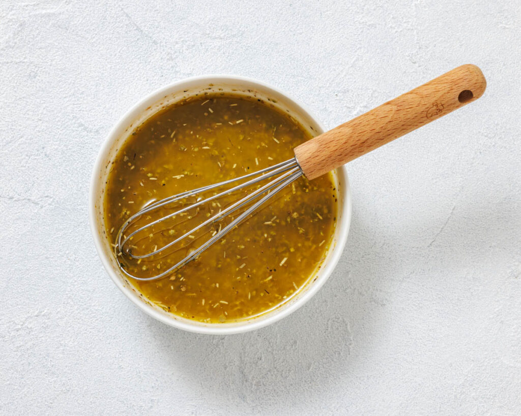 A bowl of yellow-green salad dressing with herbs, stirred with a metal whisk that has a wooden handle, on a light textured surface.