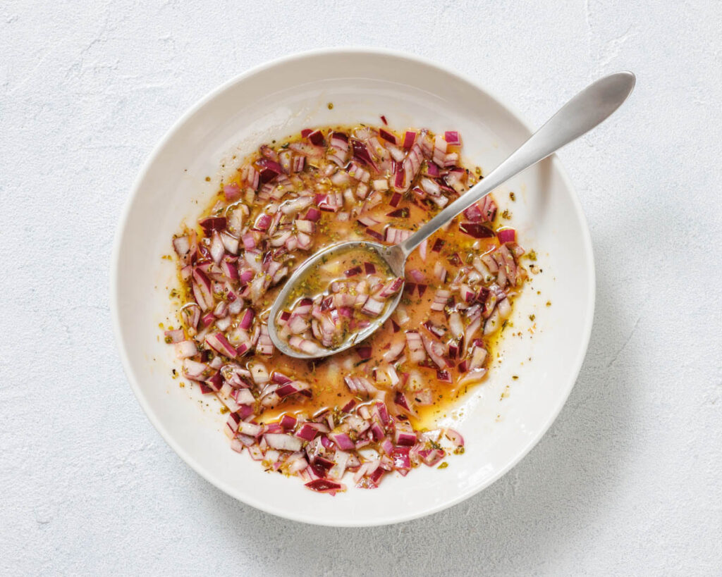 A white bowl containing a mixture of chopped red onions, oil, and herbs, with a spoon resting inside.