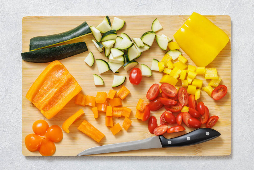 A cutting board with chopped zucchini, yellow bell pepper, orange bell pepper, cherry tomatoes, and a kitchen knife.