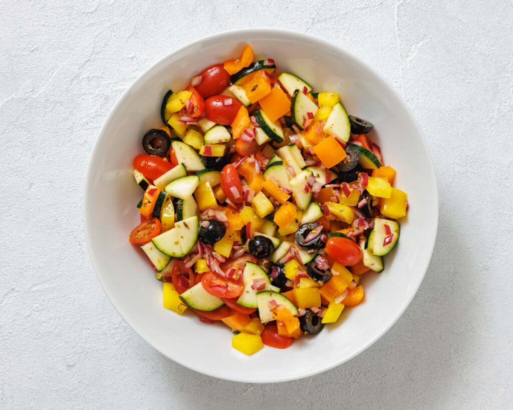 A white bowl filled with a chopped vegetable salad including tomatoes, yellow bell peppers, cucumbers, red onions, and black olives on a light gray surface.