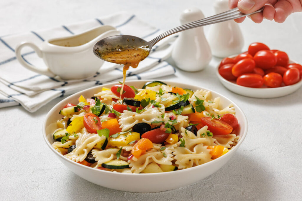 A spoon drizzles dressing over a bowl of pasta salad with cherry tomatoes, zucchini, yellow peppers, and herbs; a bowl of tomatoes and dressing are in the background.