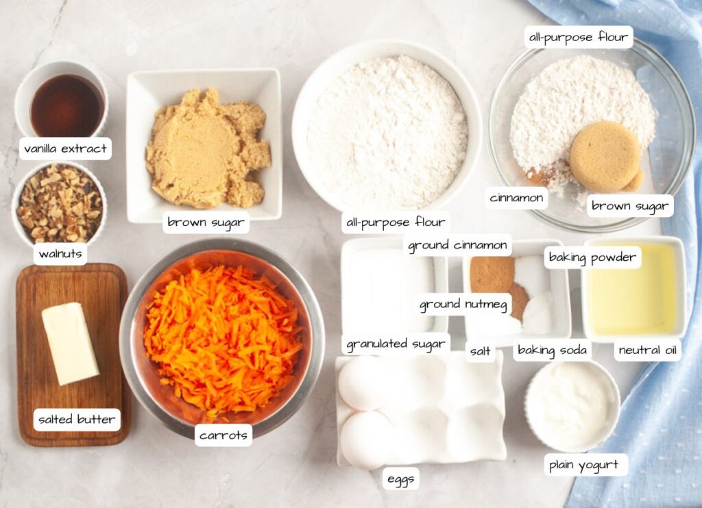 Overhead shot of labeled ingredients for carrot muffin, including carrots, flour, sugar, brown sugar, eggs, butter, oil, walnuts, vanilla, cinnamon, nutmeg, baking powder, baking soda, salt, and yogurt.