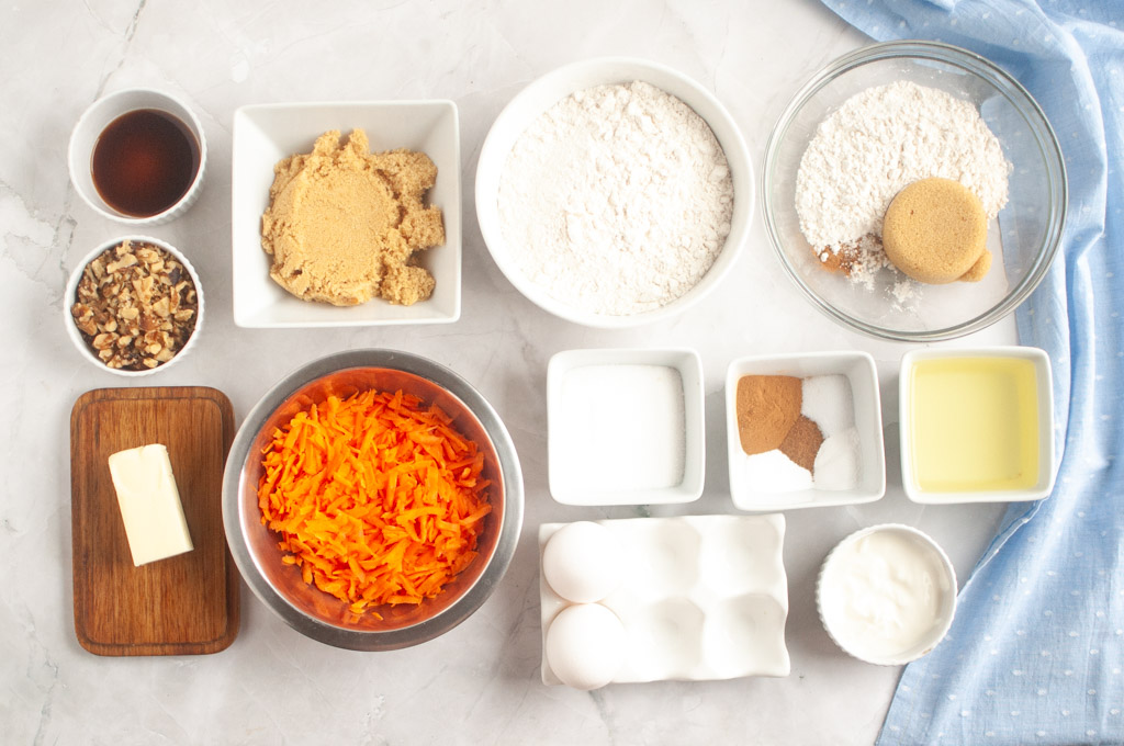 Overhead shot of ingredients for carrot muffin, including shredded carrots, flour, sugar, brown sugar, eggs, butter, oil, walnuts, vanilla, cinnamon, and baking soda.
