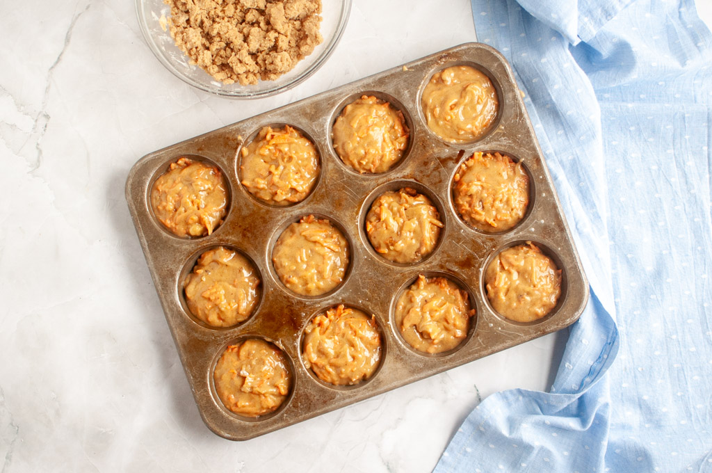Overhead shot of carrot muffin batter filled in a muffin tin, with streusel topping nearby.