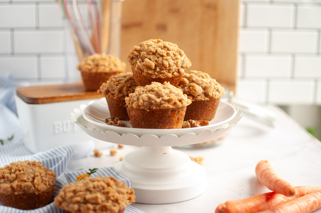 A white cake stand holds a stack of banana nut muffins topped with streusel, with more muffins and ingredients around it.