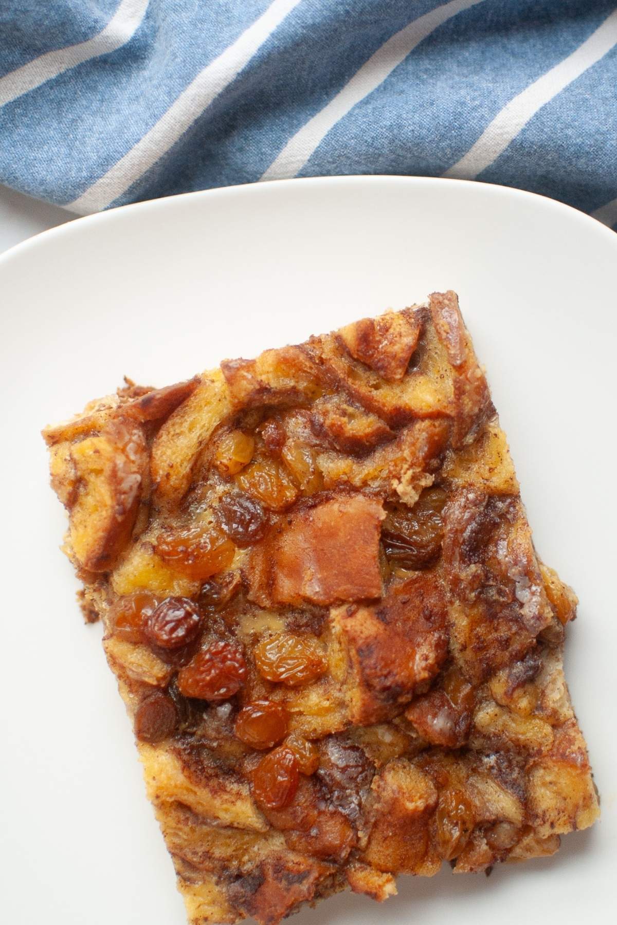 Overhead shot of a slice of golden-brown cinnamon roll casserole with visible cinnamon swirls and raisins on a white plate.