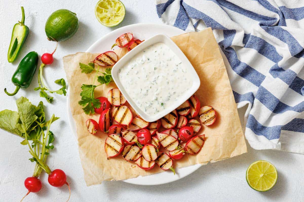 A plate with grilled radishes, a bowl of creamy dipping sauce, lime halves, jalapeño, and a striped cloth on a white surface.