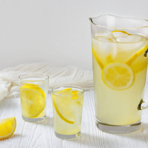 A tall glass pitcher filled with lemonade, ice, and lemon slices stands on a white wooden table, accompanied by two smaller glasses also containing lemonade and lemon. A lemon wedge is placed beside the glasses. A white cloth is draped in the background.