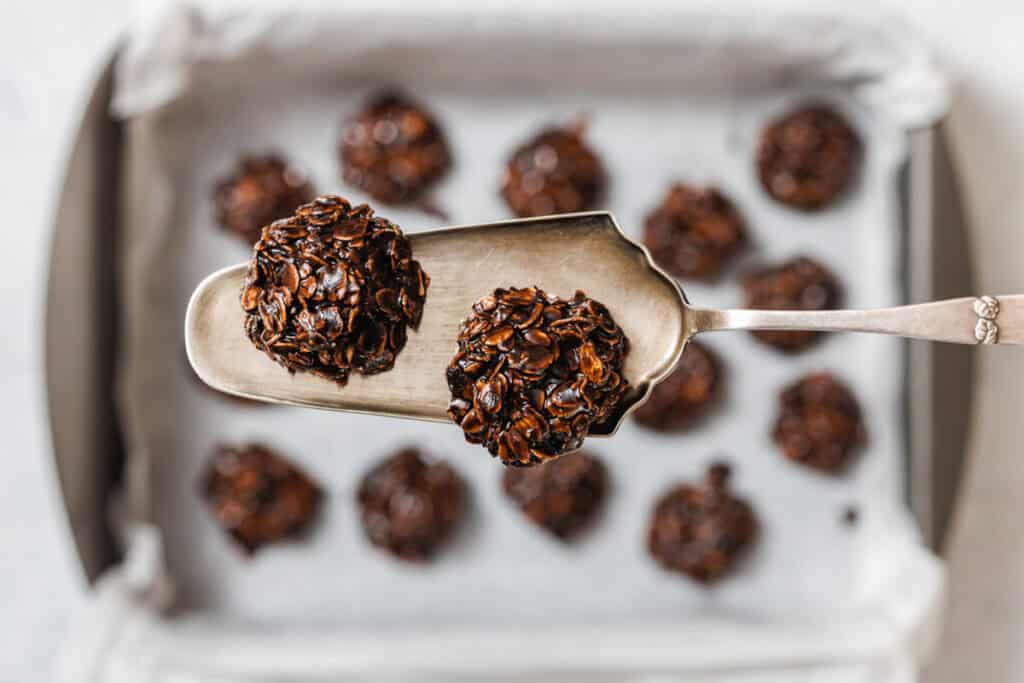 A metal spatula holds two chocolate Peanut Butter No-Bake Cookies above a baking tray lined with parchment paper and more cookies.