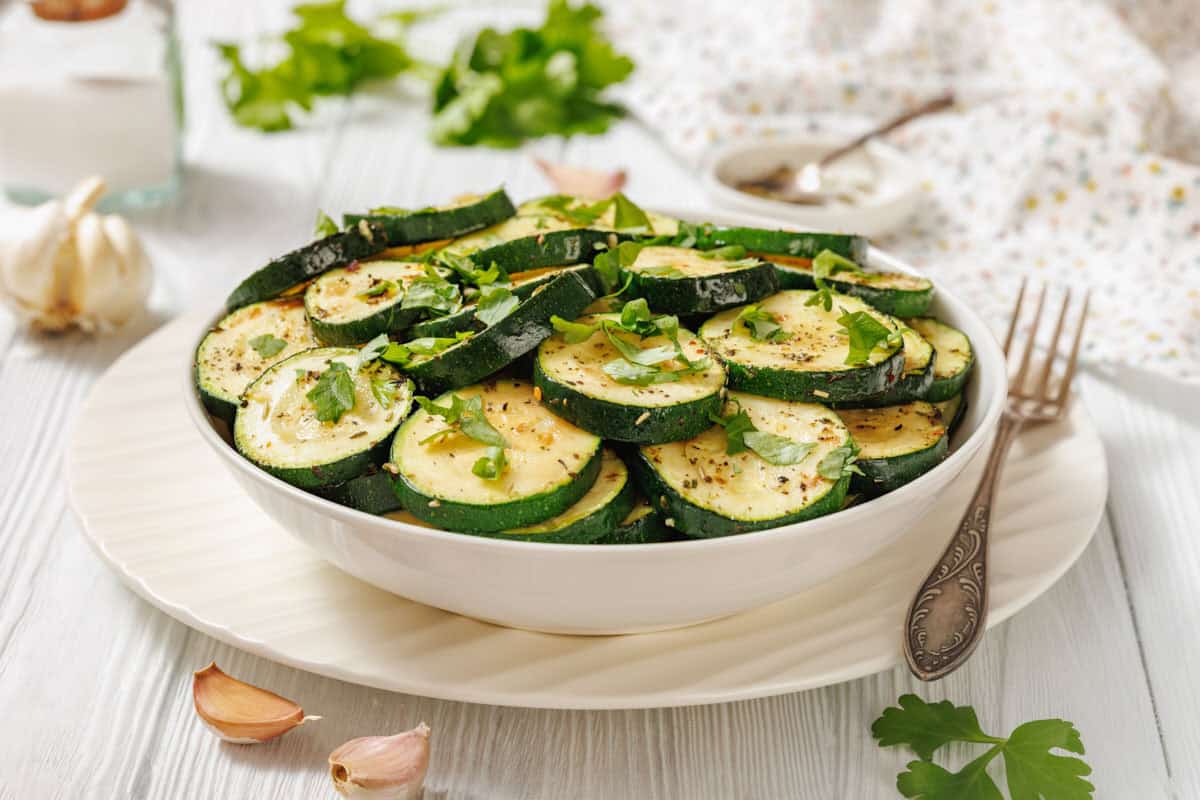 A white bowl filled with sliced Saut&eacute;ed Zucchini with Garlic and Herbs, garnished with chopped herbs, is placed on a white plate with a fork nearby.