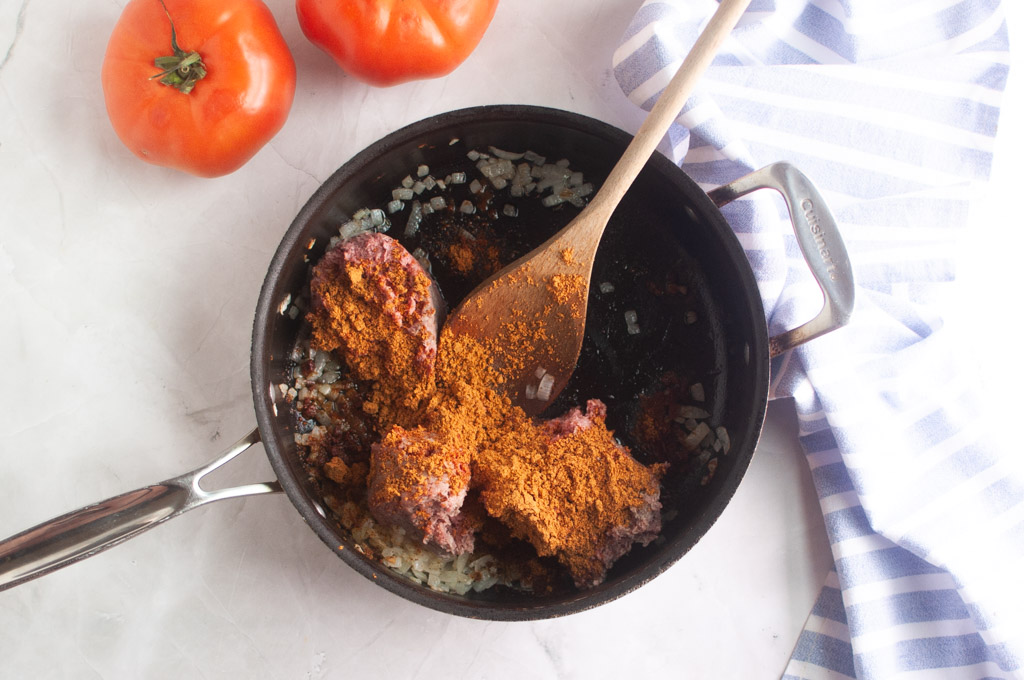 Overhead shot of ground meat, diced onions, and spices being stirred in a black skillet with a wooden spoon.