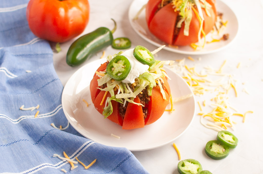 Two taco stuffed tomatoes on white plates with toppings.