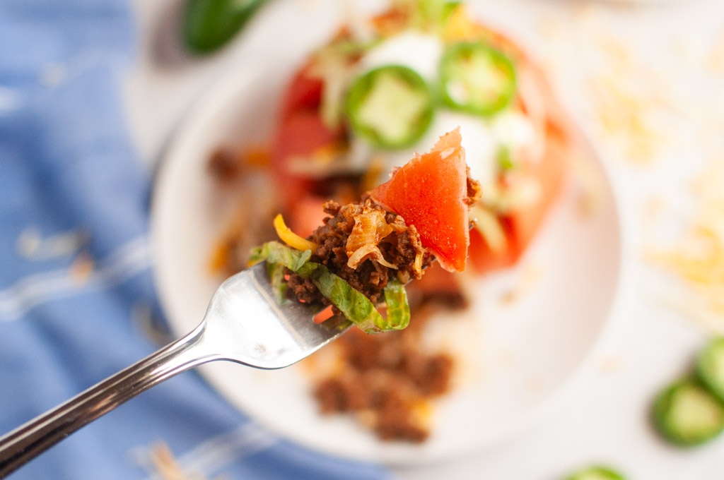 A fork holds a bite of taco stuffed tomato with meat, lettuce, and cheese.