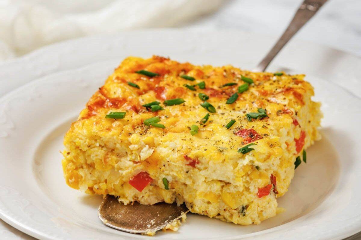 A square slice of corn pudding casserole with eggs, red bell pepper, and chives sits on a white plate with a serving spoon underneath.