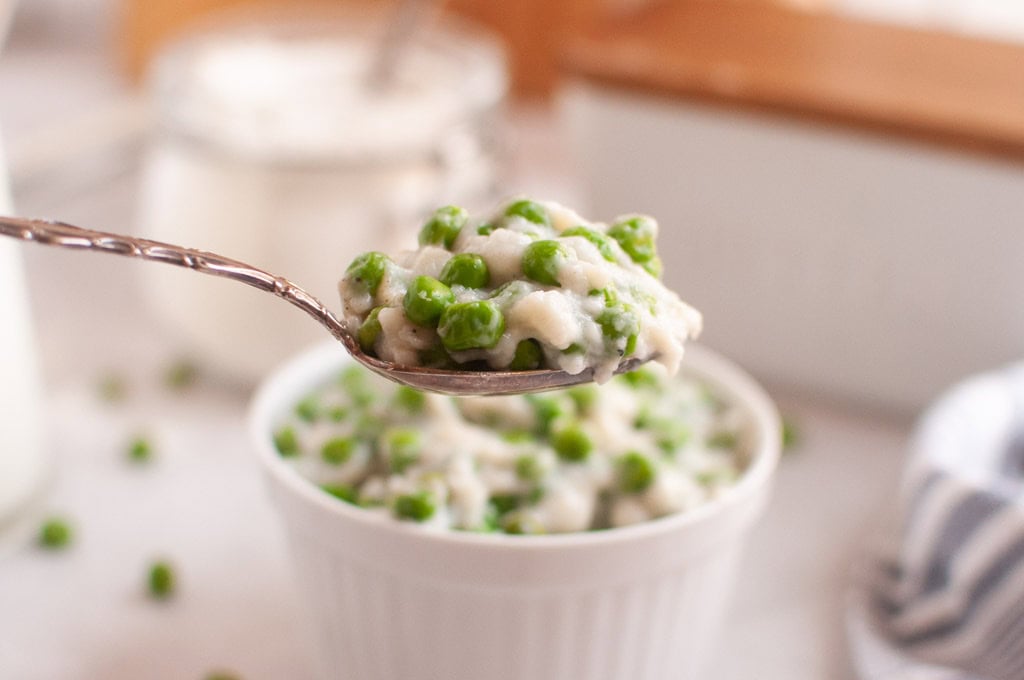 A spoonful of creamed peas is held above a white ramekin filled with the same dish, with green peas visible throughout.