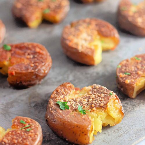 Roasted smashed potatoes topped with herbs and seasoning are arranged on a baking sheet, with one potato partially eaten to show the inside.