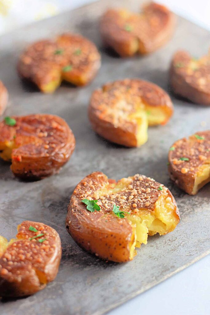 Roasted smashed potatoes topped with herbs and seasoning are arranged on a baking sheet, with one potato partially eaten to show the inside.